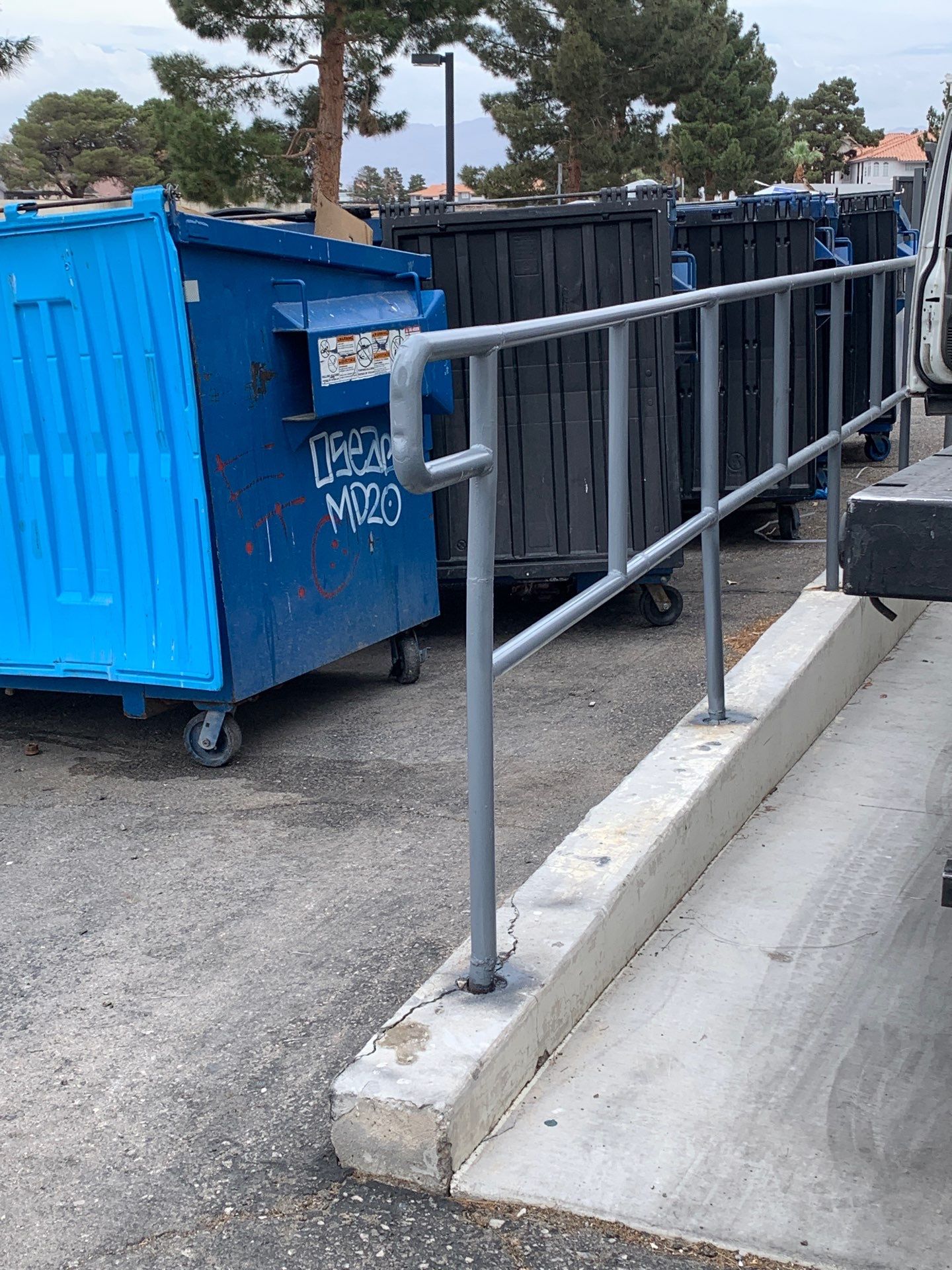 Blue dumpster beside a row of black dumpsters, with a concrete ramp and metal handrail.