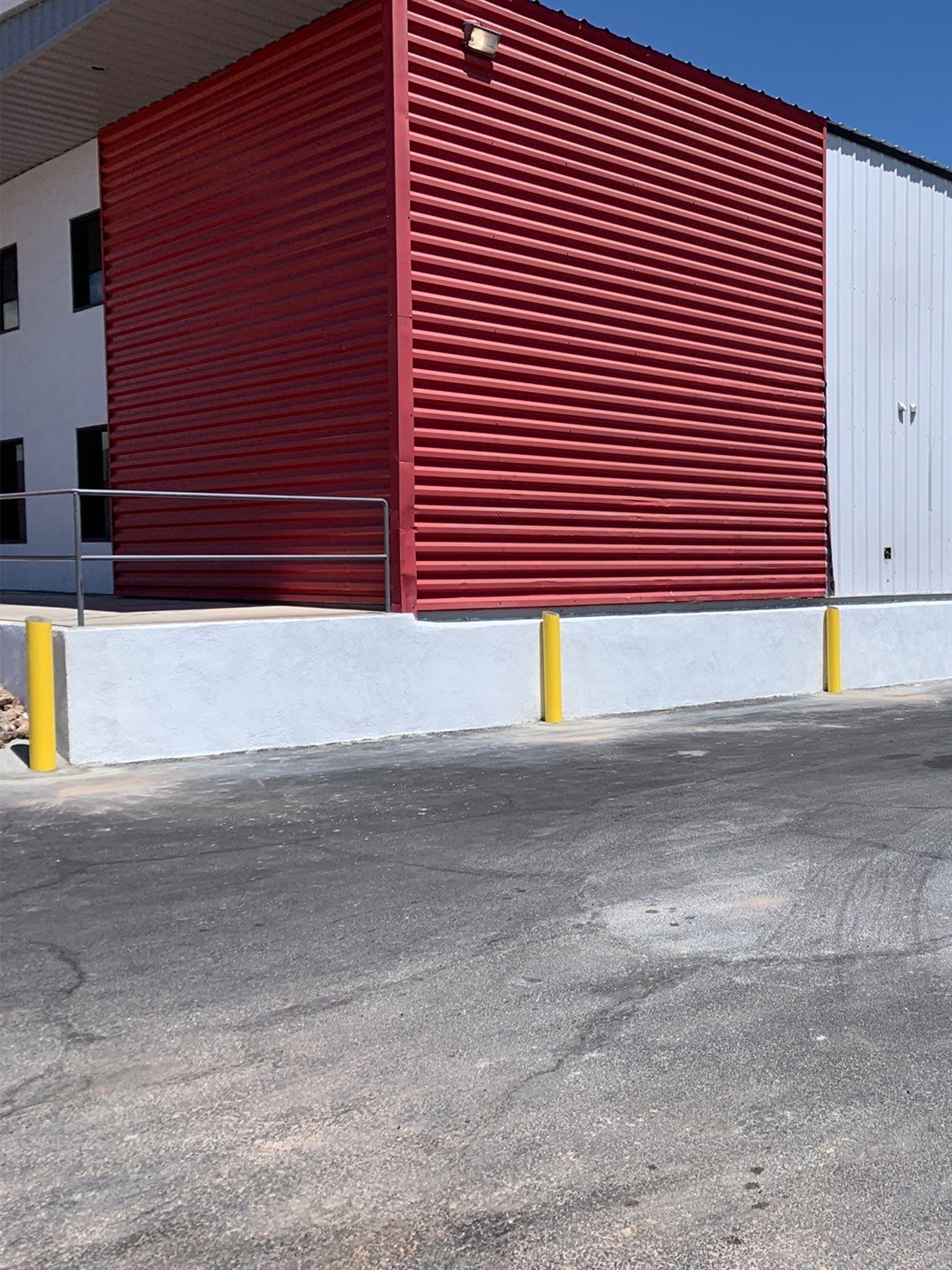 Red corrugated metal wall of a building with white trim and yellow bollards along a concrete curb.