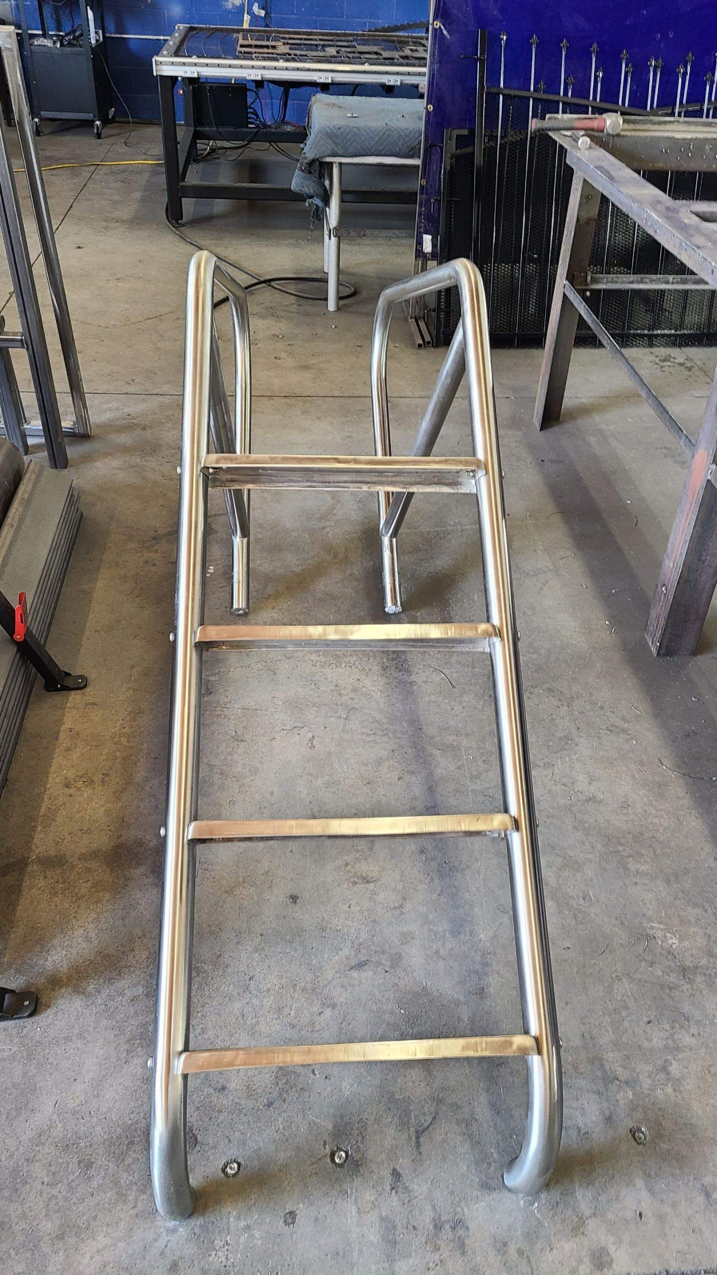 Silver metal step ladder standing on a concrete floor inside a workshop with a blue workbench in the background.