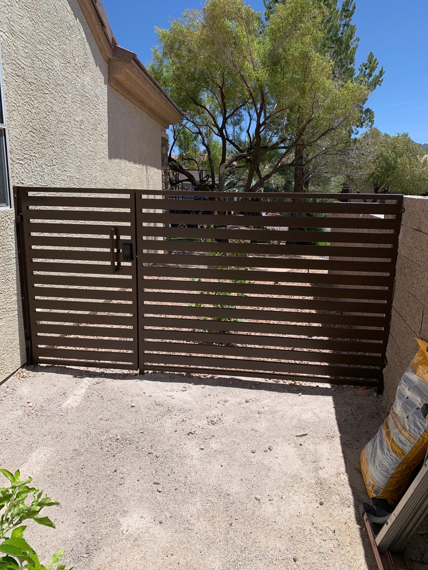 Brown horizontal-slatted gate across a concrete path in a sunny courtyard with a beige wall and plants nearby.