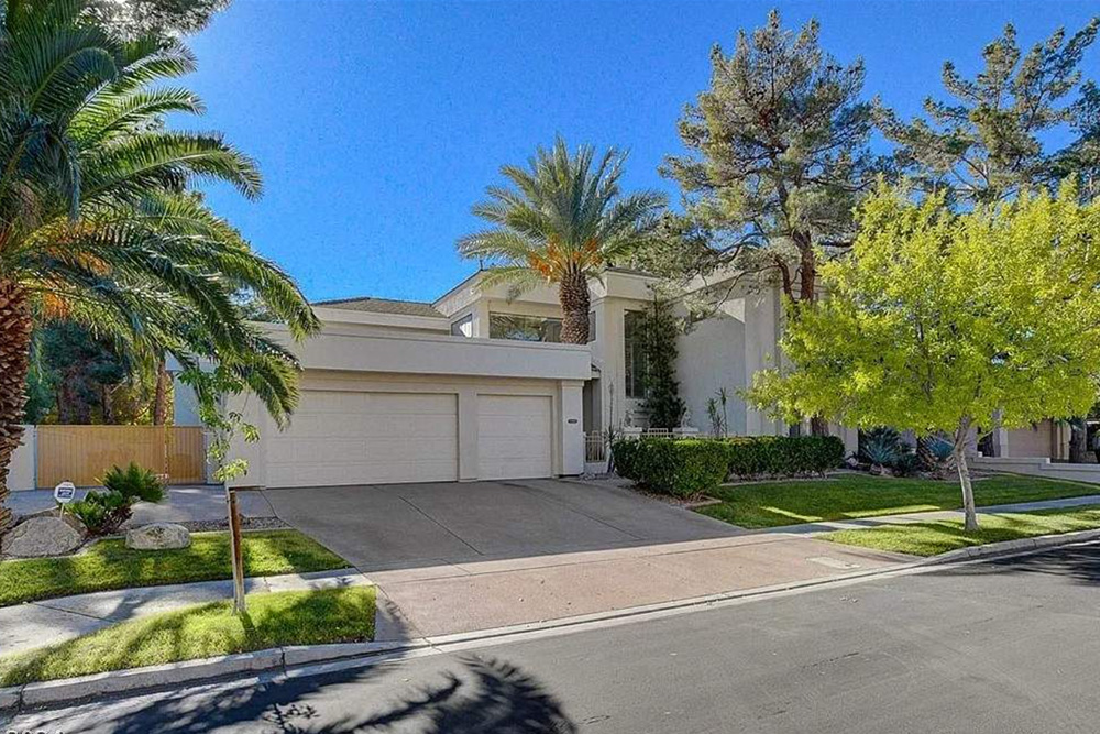 Wide shot of a modern white house with a two-car garage, manicured lawn, and palm trees under a blue sky.